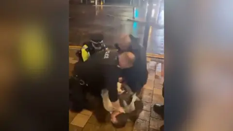 Colin Allan, in a cream blazer, is being held face down on a pavement by three police officers, one of which has his hand on the back of Mr Allan's neck