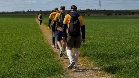 Contributed Several people are walking on a small mud path which leads between a field of green grass. Three of them are in the foreground and are wearing orange T-shirts. There are people further along the track.