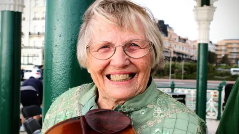 An elderly woman with short grey hair and glasses. She is wearing a light green top and smiling at the camera