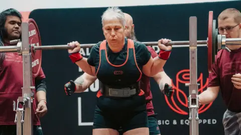 Dee Atkins-Greig in a powerlifting competition. The barbell is on her shoulders, her face shows effort.