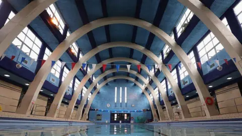 Getty Images Swimming pool with stone arches above the water. Large brown doors are visible at the far end. There are high windows and both sides and three long thin windows at the far end.