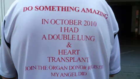 Steve Cervi A man, pictured from behind, wearing a white t-shirt. In red lettering is printed: 'Do something amazing. In October 2010 I had a double lung & heart transplant. Join the organ donor register. My Angel did."