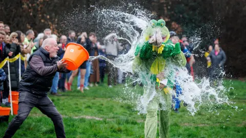 PA Media A person running along in a green frog costume is splashed by a bucket of water