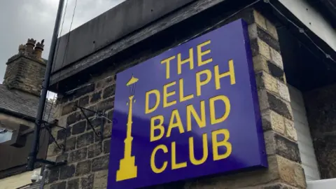 Sign reading: 'Delph Band Club' on a brick wall.