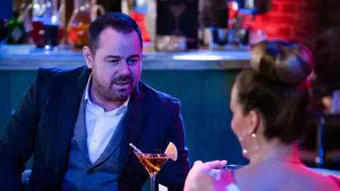 Danny Dyer melihat ke seberang meja ke arah seorang wanita di depan sebuah bar. Dia mengenakan rompi abu -abu dan jaket jas gelap dan kemeja putih. Anda dapat melihat bagian belakang kepalanya - roti pirang, anting -anting lingkaran dan atasan merah muda. Dia terlihat seperti Linda, istrinya di EastEnders, dan pengaturannya terlihat seperti salah satu bar di set EastEnders. Ada minuman jeruk di kelas koktail dengan sedotan di antara mereka. Di bar ada pitcher dari berbagai minuman berwarna. Wanita itu terlihat seperti dia tersenyum tetapi Anda tidak bisa melihat banyak wajahnya. Dia menatap matanya.