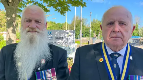 Paddy, left, is looking at the camera and has a very long grey beard. He is wearing military uniform and has a number of medals on his left lapel. Brian, right, is looking at the camera. He is bald and also wearing military dress. He has a blue and yellow ribbon around his neck and also has a number of medals on his left lapel.
