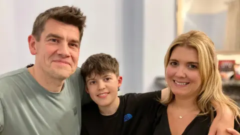 Dan Burrows, wearing a grey t-shirt, stands with his arms around his son, Lucas, wearing a black shirt and his wife Lucy, wearing a gold necklace and black top