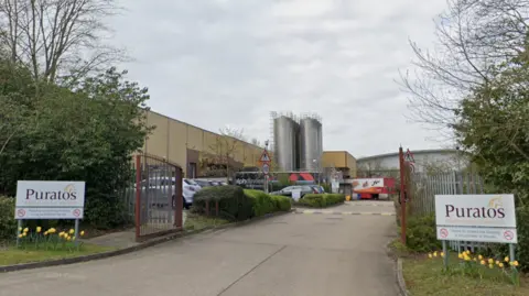 The entrance to a manufacturing site belonging to Puratos, a wholesale bakery. In the foreground is a wide access road leading through open metal security gates into the large warehouse's car park.