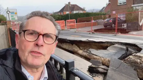 BBC reporter Adrian Harms stood next to a huge sinkhole in a road