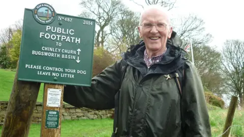 David Morton A man in a green coat wearing glasses smiling for the camera. He is in a countryside setting stood next to a green sign which reads N487 2014 PUBLIC FOOTPATH TO CHINLEY CHURN BUGSWORTH BASIN PLEASE CONTROL YOUR DOG LEAVE NO LITTER.