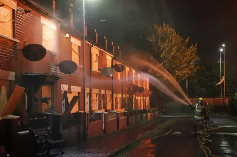 PA Media Image of two fire fighters spraying a terraced house with water, the windows are boarded up and it is dark outside, with a red glow from nearby flames