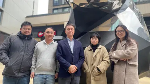 Five smartly dressed men and women stand together in front of a mirrored statue.