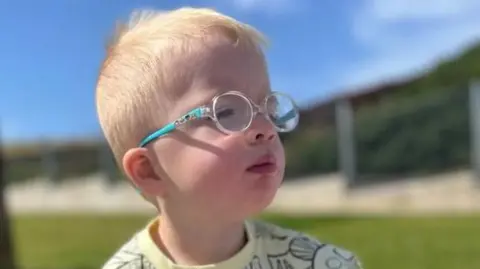 Frankie sitting on some grass, looking off to the right. He has a patterned yellow top on, short blond hair and glasses with blue arms