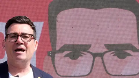 PA Andy Burnham with dark hair, dark specs, blue jacket and white T-shirt at a campaign e vent with an avatar of him in the background