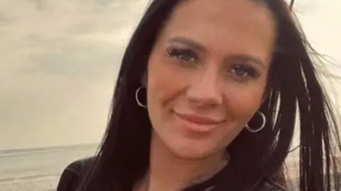 Head and shoulders image of Kiena Dawes. She is on a beach with the sea in the background, is smiling and has long brown hair