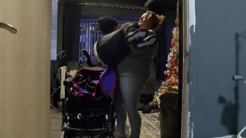 A woman wearing grey casual clothes struggles to lift a 12-year-old boy from his wheelchair inside the living room of their council house, which is decorated with a Christmas tree and lights. 