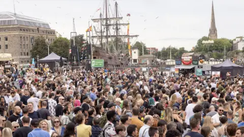 Paul Box A large crowd of people standing in Lloyd's Amphitheatre during Bristol Harbour Festival. The sky is overcast but bright, and in the background you can see a tall galleon ship moored in the harbour, with Spanish flags and bunting flying from its mast.