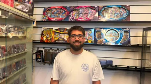 A man stands inside a trading card shop, surrounded by shelves of Pokémon boxes and a glass cabinet of cards, wearing glasses and a white T‑shirt.