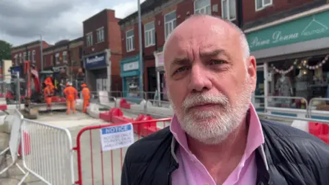 NATHAN TURVEY/BBC A picture of William Marshall, who has white hair and a white beard and is wearing a pink polo shirt and blue jacket, standing in front of some roadworks in Brighouse. There are metal barriers and workmen in the background and a row of shops. 