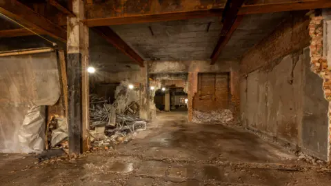 Stockton-on-Tees Borough Council A large room has been stripped to its bare red brick and concrete walls. There are bright lights coming from lightbulbs strung overhead. There is a pile of metal and brick in the back left corner.