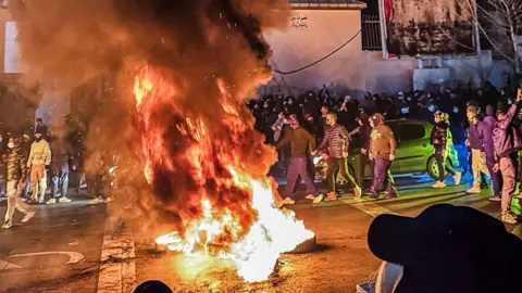 Getty Images A large fire can be seen in the middle of a road, while cars are parked beside it. There are dozens of people surrounding the flames, some of them are wearing face masks, while others are not. 