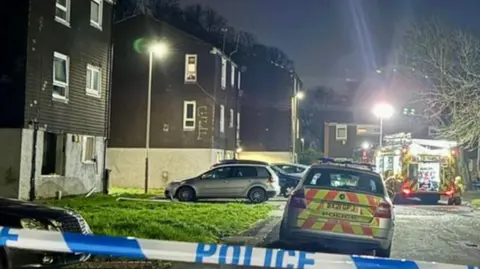 The picture shows a blown out ground floor window. There is police tape in the foreground and shows a police car and a fire engine. 