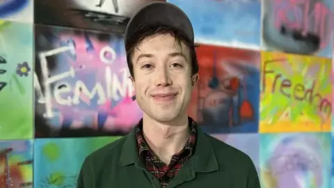 Kris Holland/BBC Dan Rainford in a baseball cap. He is wearing a green polo top with an open collar smiles at the camera. Behind him are street art designs.