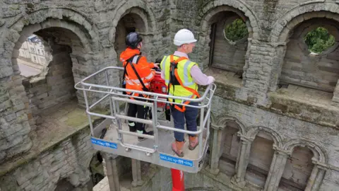 HES Two people in hard hats on a raised platform examine the stonework of Kelso Abbey