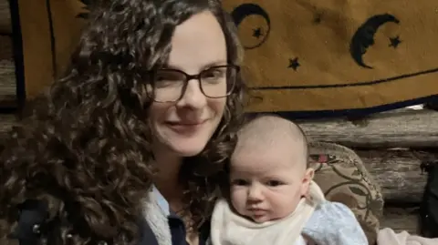 Jordan and nine-week-old Beatrice. Jordan has long black hair, a Beatrice is sat on her knee with a blue outfit on and a white bib.