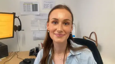 Mousumi Bakshi/BBC Head and shoulders of Heidi Brown, sitting in an office. She has long mid-brown hair pulled back from her face. She is wearing a light grey shirt. Behind her is a desk, a monitor and mouse.