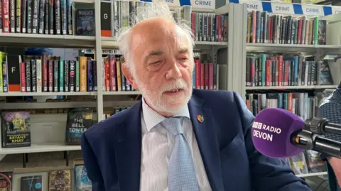 A smartly dressed man sits in front of a bookshelf in front of a microphone. He is wearing a blue blazer and blue tie.