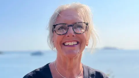 Marina Burr - A woman with wispy white hair, smiling towards the camera. She is wearing blue glasses and has a necklace with a starfish on her neck. She's wearing ablue blouse. 