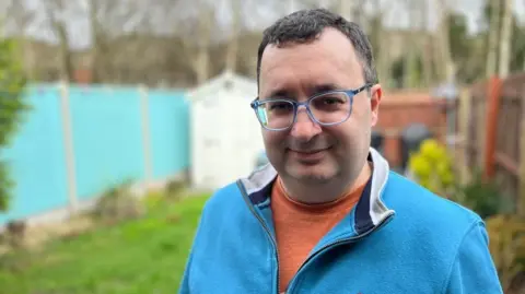 Andrew Mulloy, wearing an orange t-shirt and blue fleece jacket, is smiling at the camera while standing in his garden. 