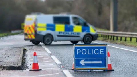 A police diversion sign on a road with two traffic cones and a police van in the background