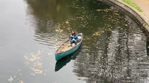 Lucy Acheson Chloe Edwards is sat paddling down Regents Canal in the Community Canoe. The Canoe is green and centre-frame in the shot.