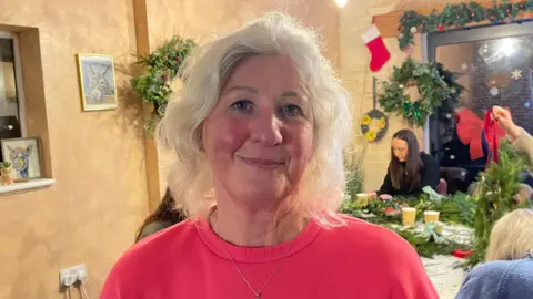 BBC/Amelia Riley A woman with shoulder-length white hair and a bright pink jumper on stands looking into the camera. Behind her, a group of people are sitting at tables making Christmas wreaths. Two completed wreaths and a garland also hang on the walls.