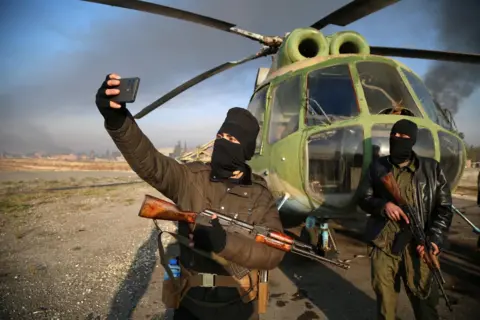 Bilal Al Hammoud/EPA-EFE Armed men take selfies at the Nayrab military airport, in Aleppo, Syria
