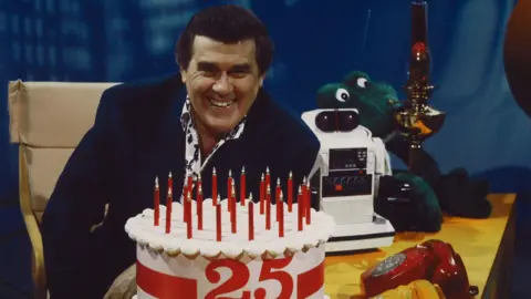 STV Glen Michael smiles while sitting at a desk. Among the items in front of him are a large fake birthday cake with 25 on it and a bright red telephone.