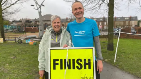 BBC/Mark Ansell Barbara and Gavin stand outdoors on a grassy area next to a paved path. They are holding a bright yellow sign with bold black text that reads “FINISH” and includes the logo and word “parkrun” at the bottom right corner. Barbara is wearing a light grey jacket, while Gavin is dressed in a bright blue T-shirt with white text that says “Capstone Foster Care” and has a long-sleeved layer underneath. In the background, there are leafless trees, a sculpture-like structure, and a row of brick houses with parked cars along the street.