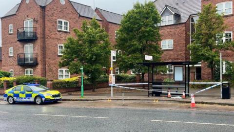 A bus stop shelter on a main road with police tape around and cones inside the cordon. A police car is parking on the left of the bus stop.