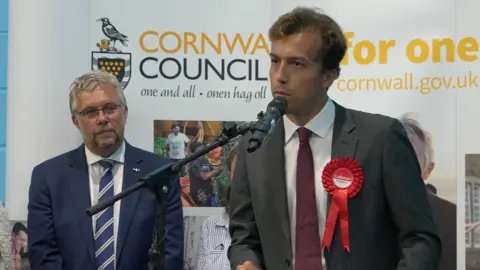 BBC Labour's Noah Law giving a speech in front of a microphone dressed in a grey suit with Steve Double behind him to the left in a blue suit and tie watching on. 