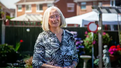 Yeti Television/BBC Cymru Wales Margaret Owen is a white woman in early old age. She has white bobbed hair, wears rimless glasses and a black and white flower-patterned blouse. She is standing outside on a sunny day. The blurred background shows housing, a dark wooden fence and colourful garden plants. She looks directly at the camera and is half-smiling.