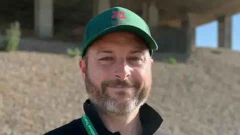 Alex Dunlop/BBC A head and shoulders picture of a man wearing a green baseball cap and a polo shirt with its collar up. He appears to be standing on a beach.