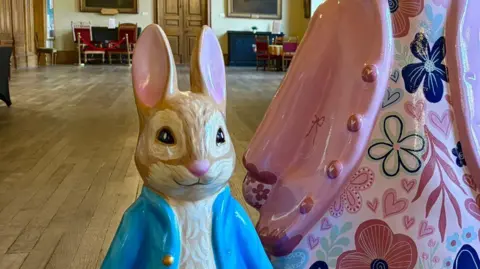 St Richard's Hospice A small Peter Rabbit sculpture, positioned alongside a larger Peter Rabbit sculpture that is mostly out of shot. The small sculpture is wearing a blue coat, the larger one is wearing a pink coat and is decorated with a floral pattern.