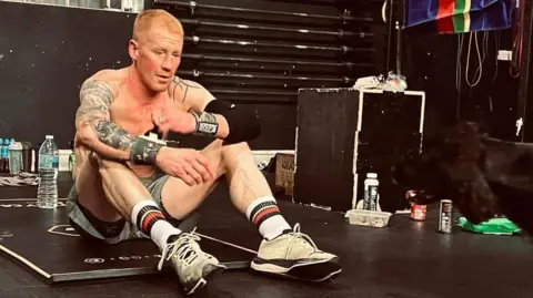 A man with short ginger hair, who is topless and wearing shorts, socks and trainers, sits on an exercise mat in a gym, looking tired.