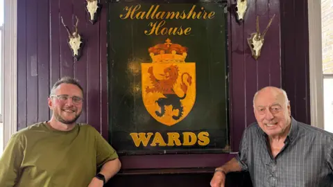 Simon Thake Two men stand smiling either side of a mantlepiece with a large pub sign hanging on the wall above them