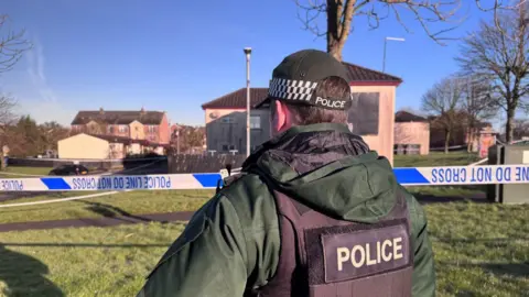 A a police officer with a dark green coat on and a black 'police' bullet proof vest and a black cap with 'police' on the back of it. In front of him are flats in a residential area and a blue and white 'police line do not cross' tape.