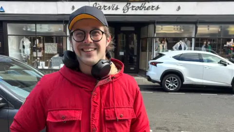A man smiling wearing headphones round his neck and a cap with a red jacket on