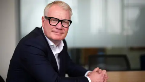 Getty Images Richard Parker's body is side on to the camera and his face is facing the camera. His hands are clasped together and he is wearing glasses. 