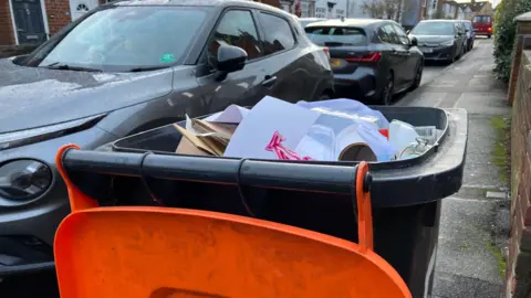 An orange-lidded bin with the lid open to reveal that it is full of paper and cardboard waste. It is standing on a pavement with cars parked alongside.